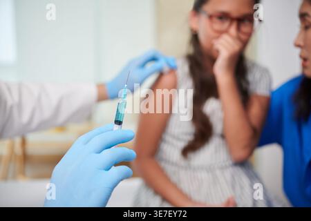Mixed race mother hugging scared Asian daughter looking at caucasian male doctor holding syringe with vaccine in clinic Stock Photo