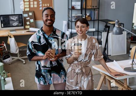Mixed-race male and female architects holding coffee cups standing near table with floor plans in office Stock Photo