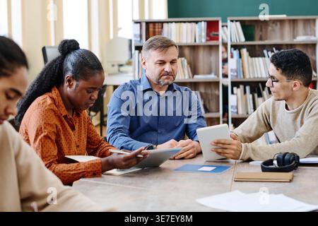 Group of immigrants doing activities on tablets while sitting at table with caucasian male instructor guiding them during English language class in... Stock Photo