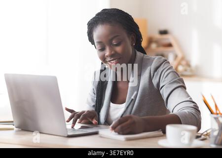 Black woman using laptop and writing in notebook sitting at table in the office during daytime Stock Photo