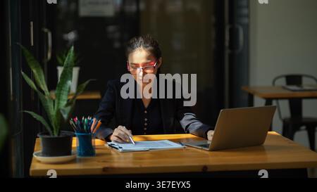 Asian woman checking documents while using laptop sitting at table in office Stock Photo