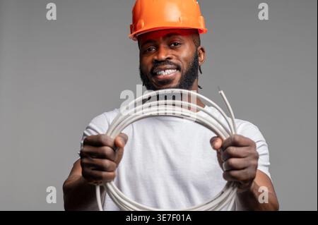 Smiling Black man wearing orange hard hat holding roll wire standing against gray background Stock Photo
