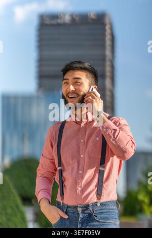 Smiling Asian bearded man having phone call standing near buildings in city  on sunny day Stock Photo