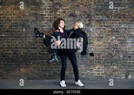 Cheerful Mixed race man with curly hair carrying caucasian girlfriend and looking at each other while standing against brick wall on street Stock Photo