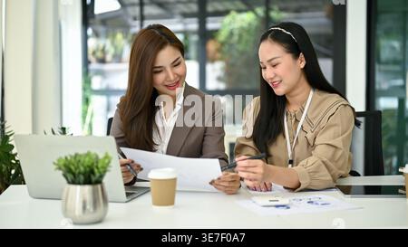 Smiling Asian female accountants working and analyzing financial data reports while sitting together at table in office Stock Photo