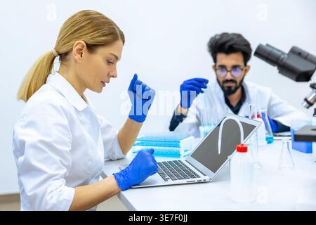 Indian male and caucasian female scientists wearing lab coats and latex gloves using microscope and laptop doing sample analysis together in labora... Stock Photo