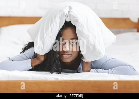 Black woman with curly hair covering head with pillow lying down in bed at home Stock Photo