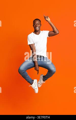 African american millennial man posing on orange studio background. Happy Black guy jumping up with tucked legs, showing up and down gesture, full lem Stock Photo