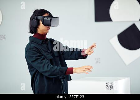 Asian man wearing VR headset experiencing virtual reality while standing in art gallery Stock Photo