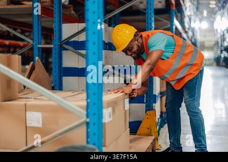 Black male worker wearing hard hat and safety vest scanning boxes using barcode scanner during work in warehouse Stock Photo