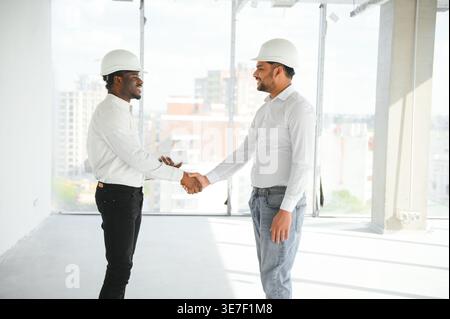 Diverse group of male engineers wearing hard hats looking at each other and shaking hands standing on construction site Stock Photo