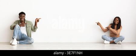 Black couple pointing fingers at each other while sitting on floor against white wall Stock Photo