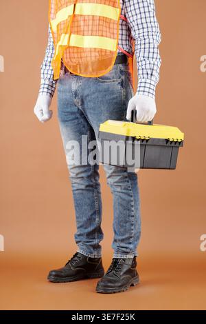 Cropped image of Asian male construction worker in jeans safety reflective vest safety shoes and protective gloves holding toolbox and standing aga... Stock Photo