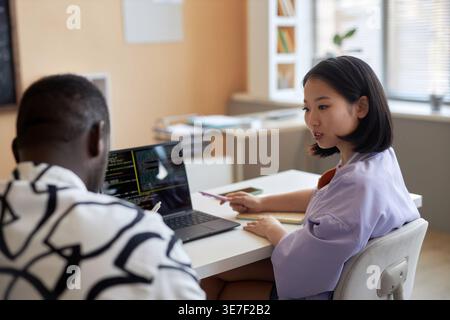 Asian female student talking to Black male classmate discussing project while sitting at desk with laptop in classroom Stock Photo