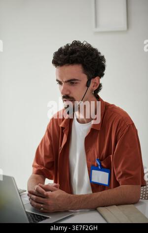 Mixed-race male office worker with clasped hands wearing headset sitting at table with laptop in office Stock Photo