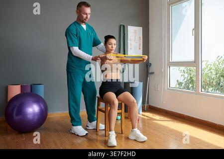 Asian female using stretch resistance band doing therapy assisted by caucasian male therapist sitting on chair in clinic Stock Photo