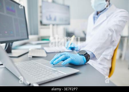 Black male scientist wearing lab coat and mask using laptop and taking notes on notebook while sitting at desk in laboratory Stock Photo