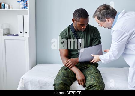 Black man with injured arm sitting on medical bed next to caucasian male doctor checking his arm cast in clinic Stock Photo
