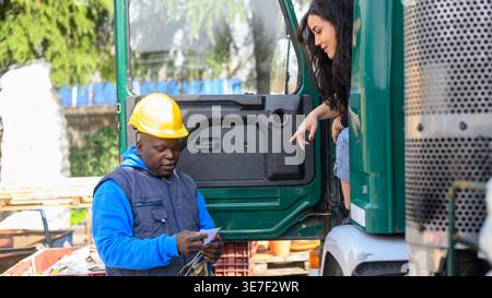 Serious Hispanic female truck driver sitting in the truck talking to Black man wearing blue vest and yellow hardhat standing at construction site Stock Photo