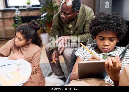 Black boy writing notes lying on sofa in front of Black grandfather and sister doing homework while sitting at table in living room at home Stock Photo