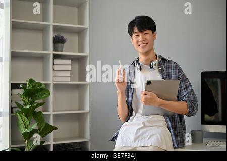 Smiling Asian male with headphones on neck holding tablet and stylus pen while standing near computer at home Stock Photo