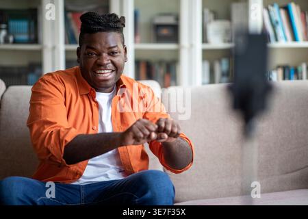 Cheerful Black man with braids recording video sitting on sofa in living room at home Stock Photo