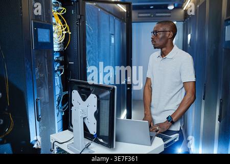 Black male IT engineer with eyeglasses looking at cables standing at table with laptop in server room in data center Stock Photo