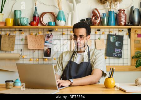 Black craftsman working on laptop sitting at table in front of porcelain tableware and sketches in workshop Stock Photo