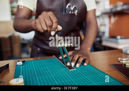 Hands of Black woman Lifestyleisan holding leather compass cutting handmade belt placed on board standing in workshop Stock Photo