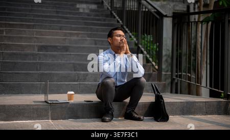 Asian businessman with praying hands looking up sitting near opened laptop, coffee, and briefcase on building stairs in city during daytime Stock Photo