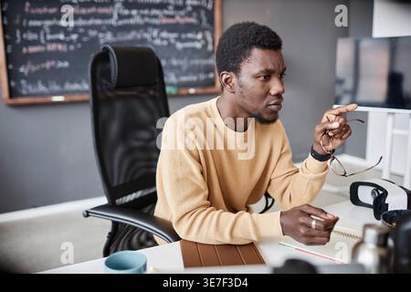 Black male programmer pointing finger and holding eyeglasses sitting at table with notebook in office Stock Photo