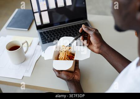 Cropped image of Black male IT specialist eating takeout noodles while sitting at table with laptop in office Stock Photo