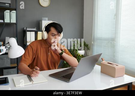 Asian male software developer wearing headphones leaning chin on hand looking at laptop while writing notes in notebook sitting at desk in office Stock Photo