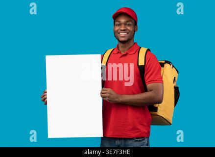 Black delivery man wearing cap carrying thermo backpack holding white blank board standing against blue background in studio Stock Photo