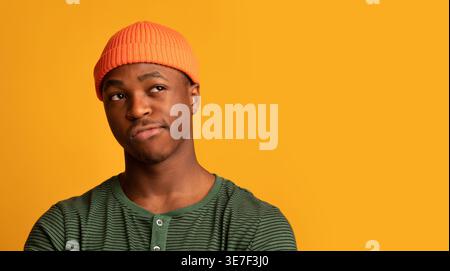 Doubtful Black man in beanie looking away while standing against yellow background in studio Stock Photo
