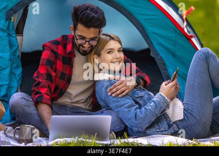 Indian man with eyeglasses wearing red checkered long sleeves using laptop while sitting with arm around caucasian female friend at tent in forest Stock Photo