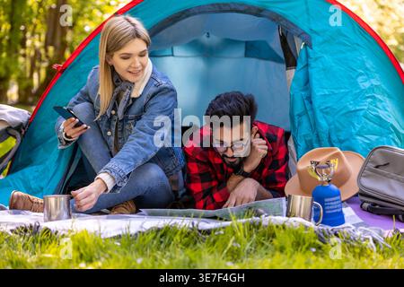 Indian man with eyeglasses wearing red checkered long sleeves checking map while lying beside caucasian female friend at tent in forest Stock Photo