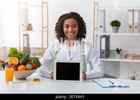 Black female nutritionist with tape measure around her neck holding tablet with blank screen while sitting at table with bowl of fruits and vegetab... Stock Photo