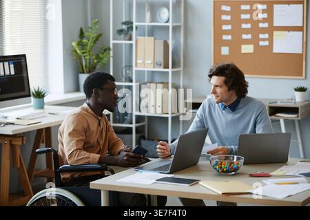 Disabled Black male IT specialist discusses work with male colleague while sitting on wheelchair near table with laptop in office Stock Photo