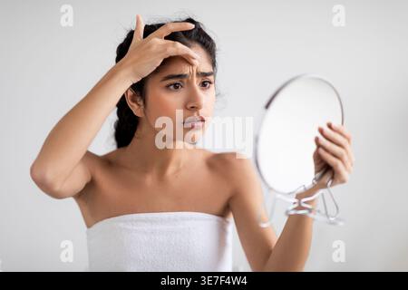 Asian woman with braids wrapped in towel holding mirror and checking wrinkles on her face standing against white wall at home Stock Photo