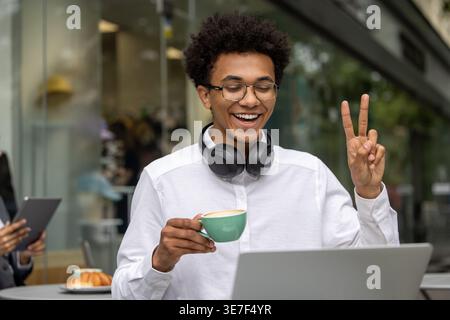 Black man wearing eyeglasses and headphones on neck doing peace sign drinking coffee and having video call on laptop while sitting at table in outd... Stock Photo