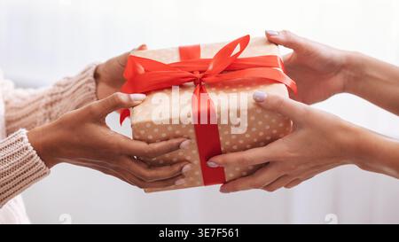 Hands of Black woman receiving gift box with red ribbon from female friend indoors Stock Photo