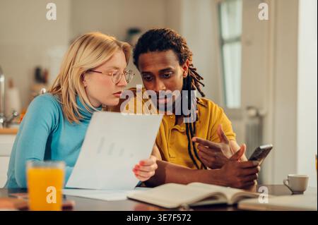 Serious Mixed race man with locks and caucasian girlfriend holding documents and smartphone having conversation sitting at table at home Stock Photo