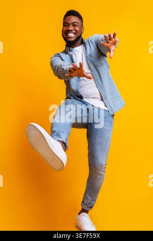 Cheerful Black man dancing and extending his hands forward against yellow background in studio Stock Photo