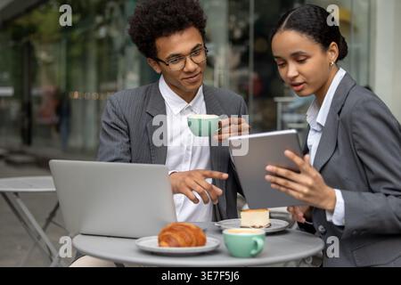 Black male and female business pLifestyleners discussing project details holding cup of coffee and using tablet while sitting together at table with past... Stock Photo