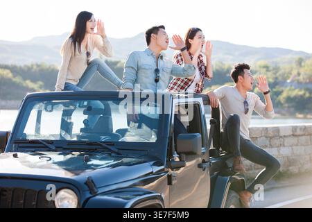 Cheerful Chinese friends shouting with cupped hands having fun together and looking way while sitting on jeep parked on road near sea wall during d... Stock Photo