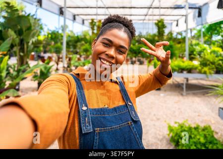 Hispanic female gardener in denim overalls showing peace sign and taking selfie while standing in greenhouse Stock Photo