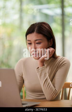 Asian woman using laptop leaning face on hand sitting at table at home Stock Photo