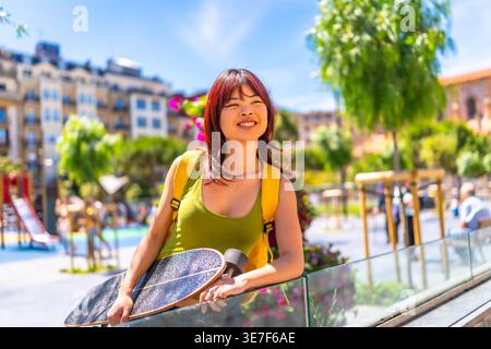 Asian woman with yellow backpack and skateboard looking away standing and leaning arm on glass railing in city on sunny day Stock Photo