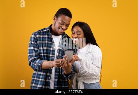 Black couple using smartphone together while standing against yellow background in studio Stock Photo